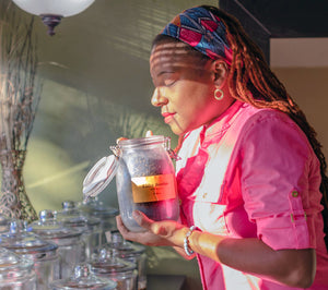 clinical herbalist in jacksonville, florida standing in an apothecary holding a glass jars of dried elderberries, sunlight shining on her face, she looks beautiful, peaceful, warm loved. She is wearing a matching head scarf and pink shirt, chakra bracelet